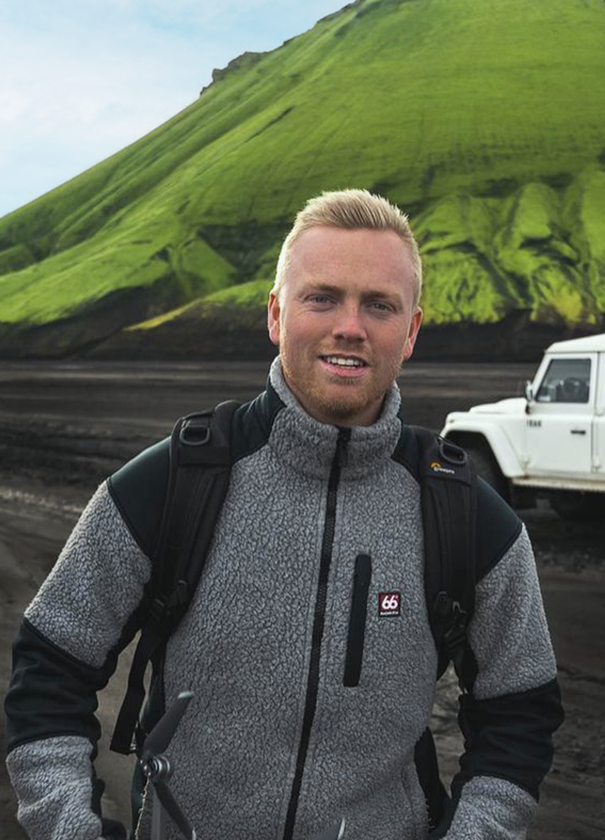 Garðar Ólafsson在一片黑沙滩上漫步，背后是一辆租来的汽车，他面带微笑，附近是碎石路。