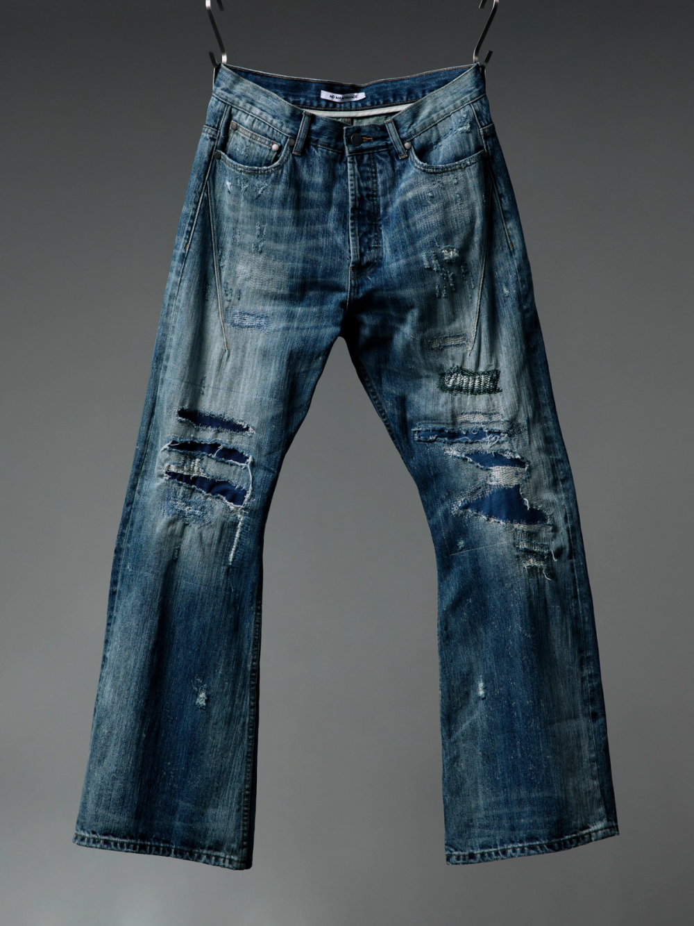 Distressed blue jeans with rips and patches hanging against a gray background.