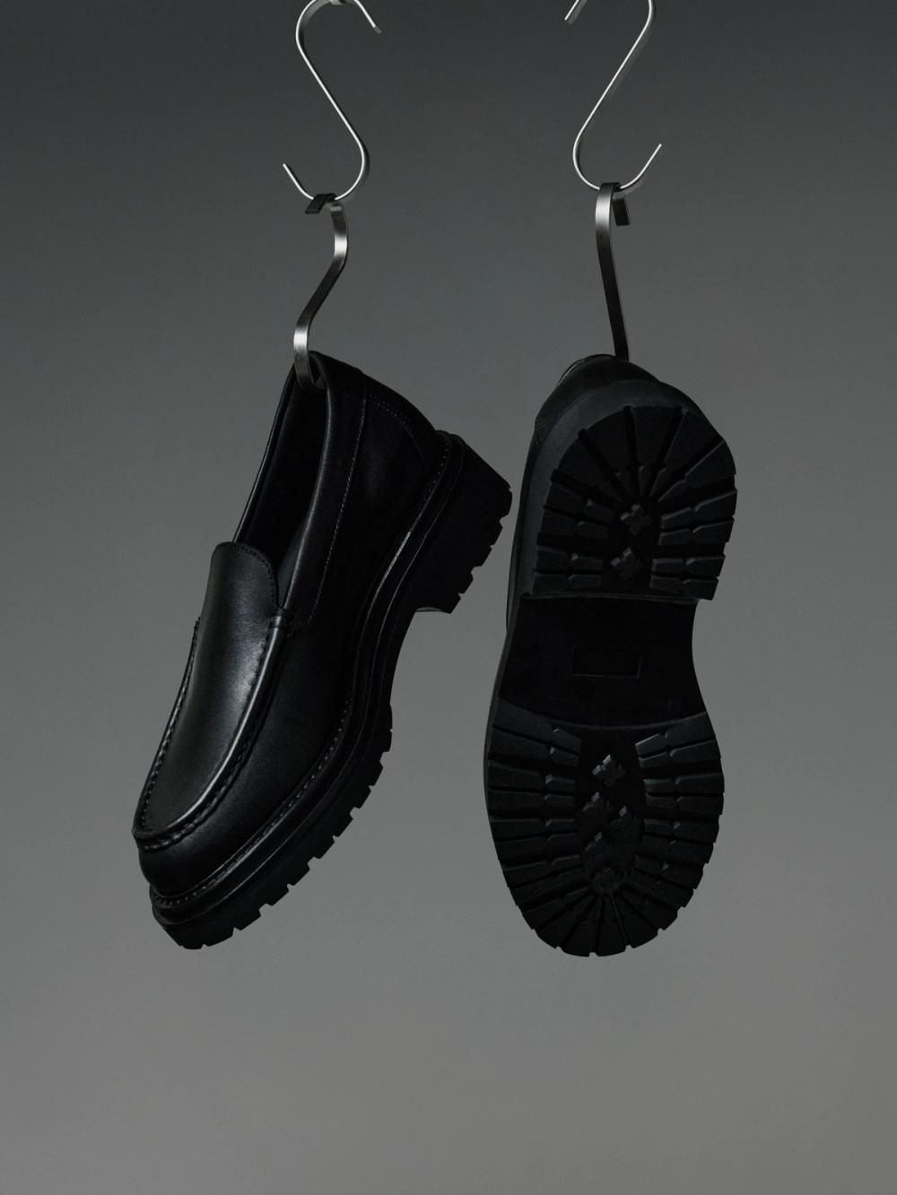 Two black chunky loafers hanging from S-hooks against a gray background.