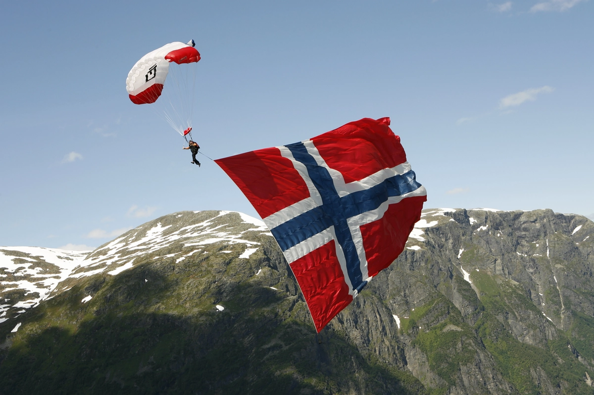 A person paragliding in the mountains with a large Norwegian flag hanging at his back