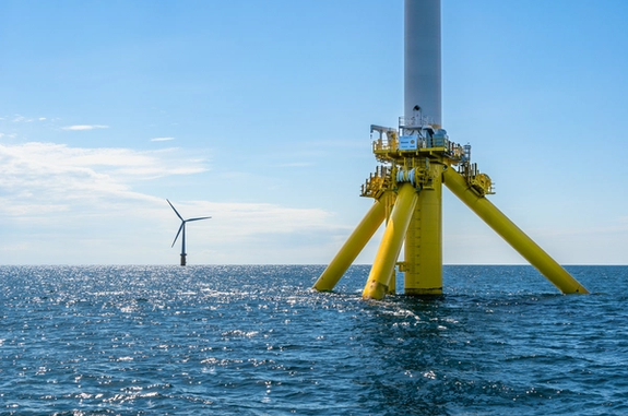 A yellow wind turbine foundation in the ocean in the front and a wind turbine in the background