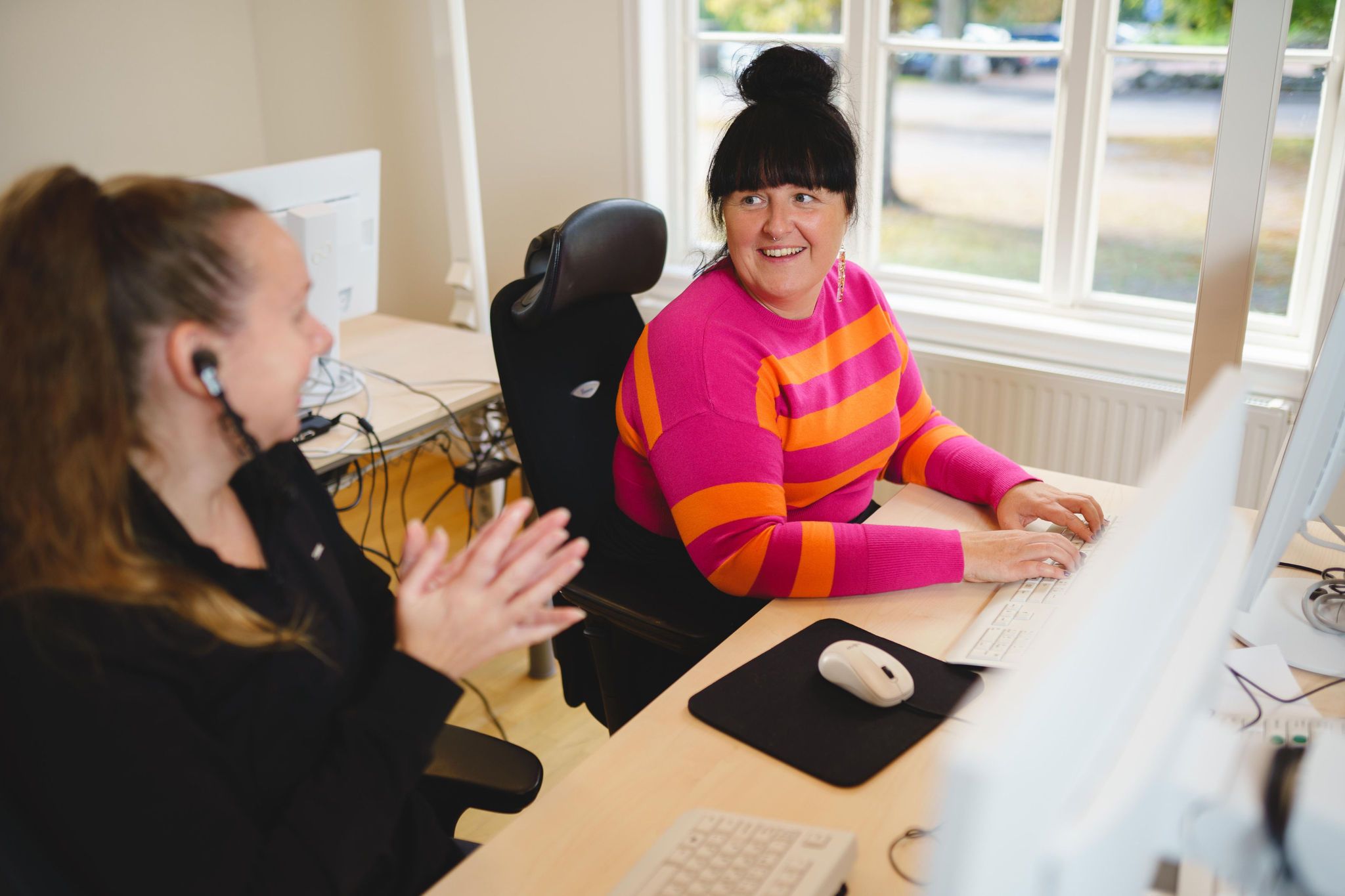 Två kvinnor sitter framför varsin dator och pratar med varandra. 