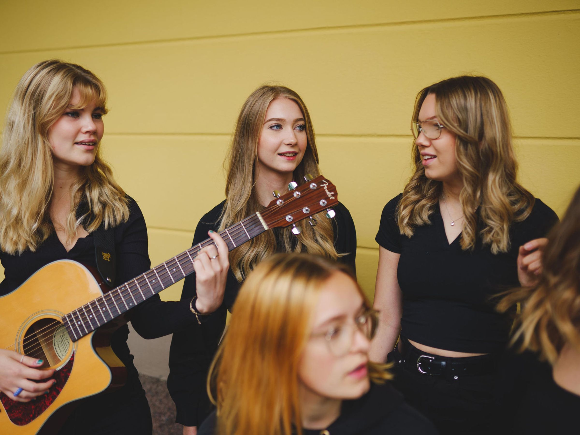Fyra svartklädda tjejer pratar med varandra. En av dem håller i en gitarr.