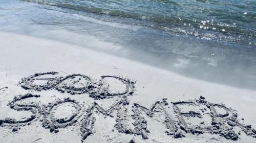 Bilde som viser teksten "god sommer" skrevet i sanden på en strand.