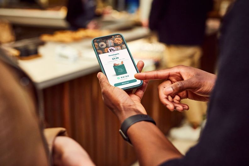 Eine Person hält ein Telefon mit einer Too Good To Go Tüte mit Brot in der Hand