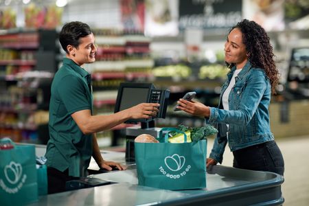 Smiling cashier helping a customer pay with her phone at a supermarket checkout, with groceries in a "Too Good To Go" bag.