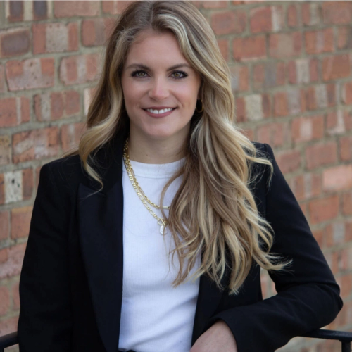 a woman wearing a black jacket and a gold necklace smiles in front of a brick wall