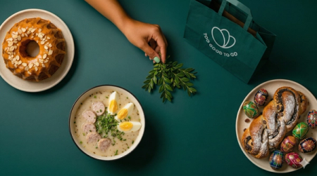 Overhead view of Easter foods: cake, soup, pastry with decorated eggs, a 'Too Good To Go' bag, and a hand arranging greenery on a green surface.