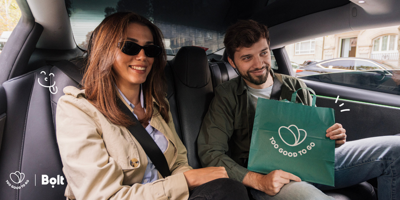 A smiling man and woman in a car, with the man holding a green "Too Good To Go" bag.