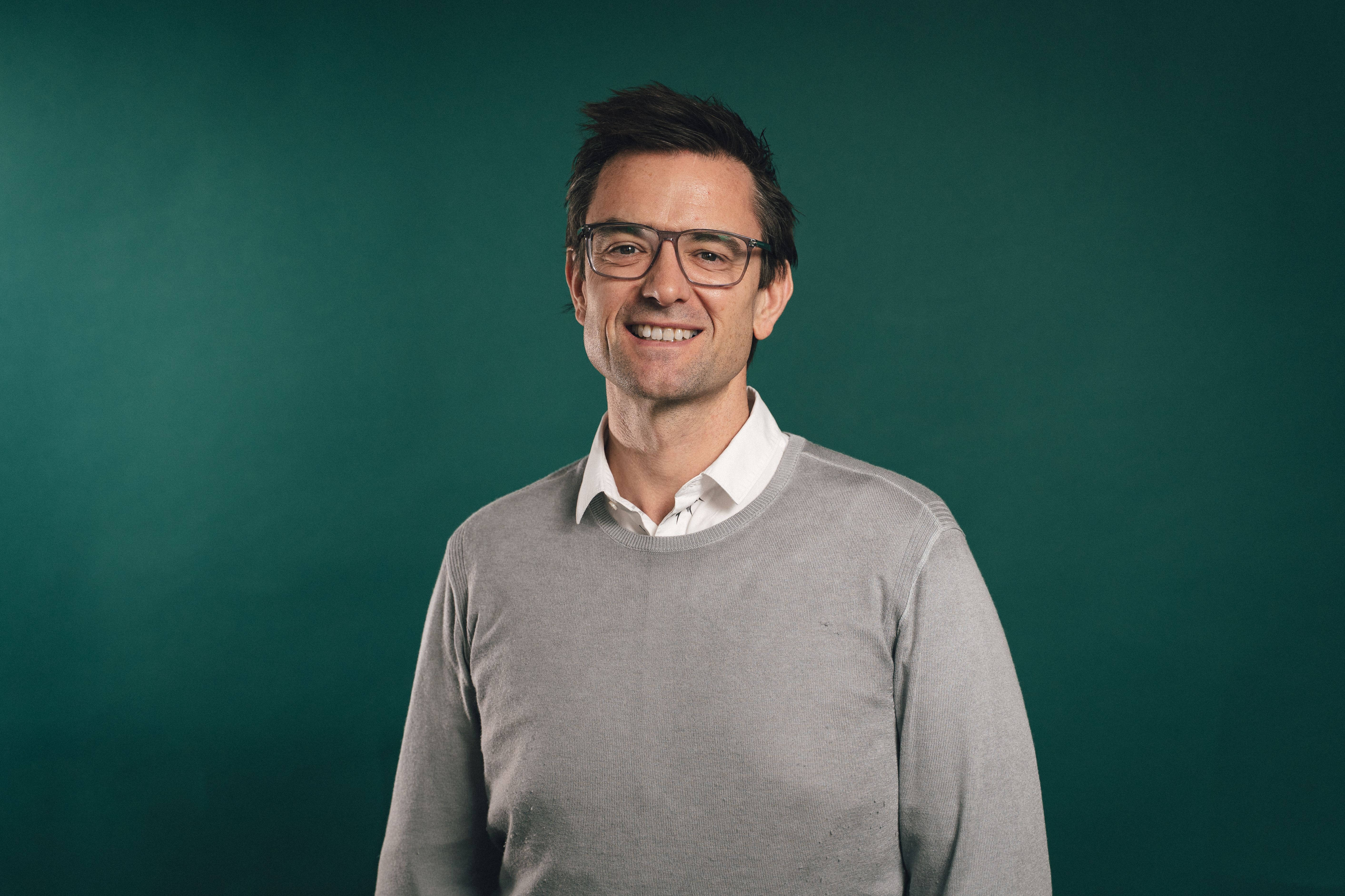 A man with a white shirt, and a gray sweater smiles against a dark green background.