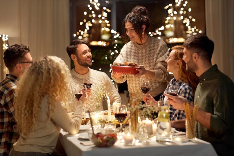 A woman serves food to smiling friends at a festive dinner party with candles and lights.