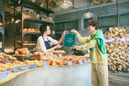 A baker hands a green shopping bag to a customer in a brightly lit bakery filled with various baked goods.