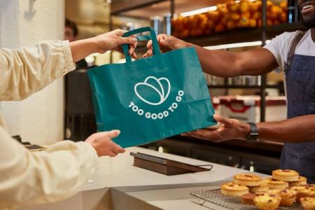 A smiling employee hands a green "Too Good To Go" bag to a customer over a counter with pastries.