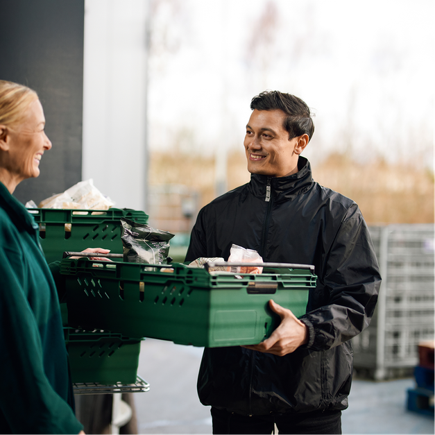 A smiling man holds a green crate of food, facing a smiling woman with more crates nearby.