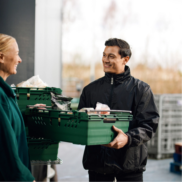 A smiling man holds a green crate of food, facing a smiling woman with more crates nearby.