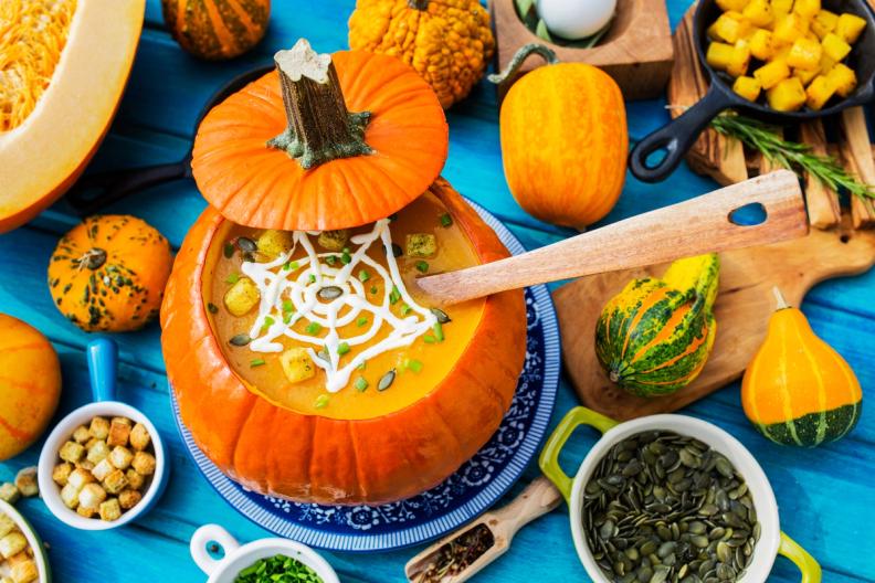 Pumpkin soup with a spiderweb design, served in a carved pumpkin, surrounded by gourds and pumpkin pieces on a blue table.