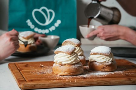 Three cream-filled semla buns on a board, with hands eating one and pouring coffee, and a Too Good To Go bag in the background.