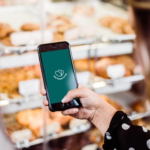 una donna tiene in mano un cellulare con il logo Too Good To Go, davanti a una vetrina di pasticcini