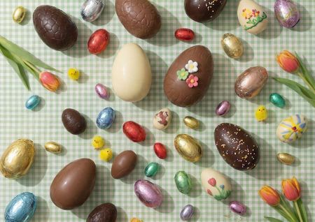 An assortment of chocolate and decorated Easter eggs, some foil-wrapped, with chick figurines and tulips on a green gingham tablecloth.