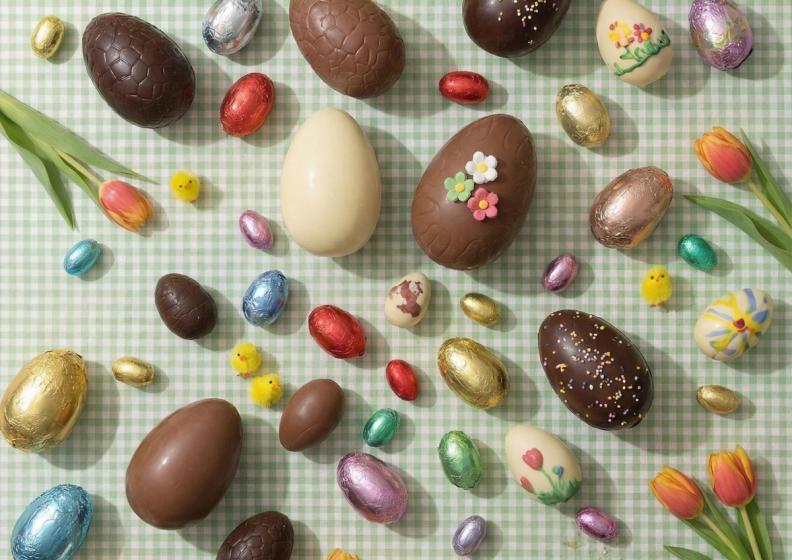 An assortment of chocolate and decorated Easter eggs, some foil-wrapped, with chick figurines and tulips on a green gingham tablecloth.