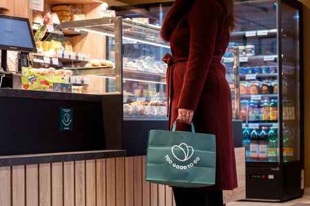 A person in a red coat holds a green "Too Good To Go" bag in a food shop.