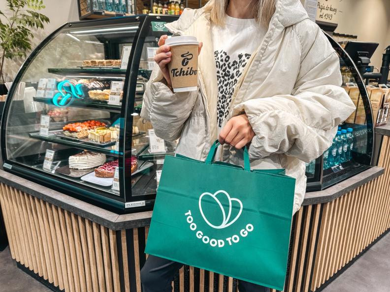 A person holds a Tchibo coffee cup and a green "Too Good To Go" bag in a cafe with a display case of pastries.