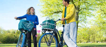 Smiling man and woman with Swapfiets bikes and Too Good To Go bags in a sunny park, with pink hearts between them.