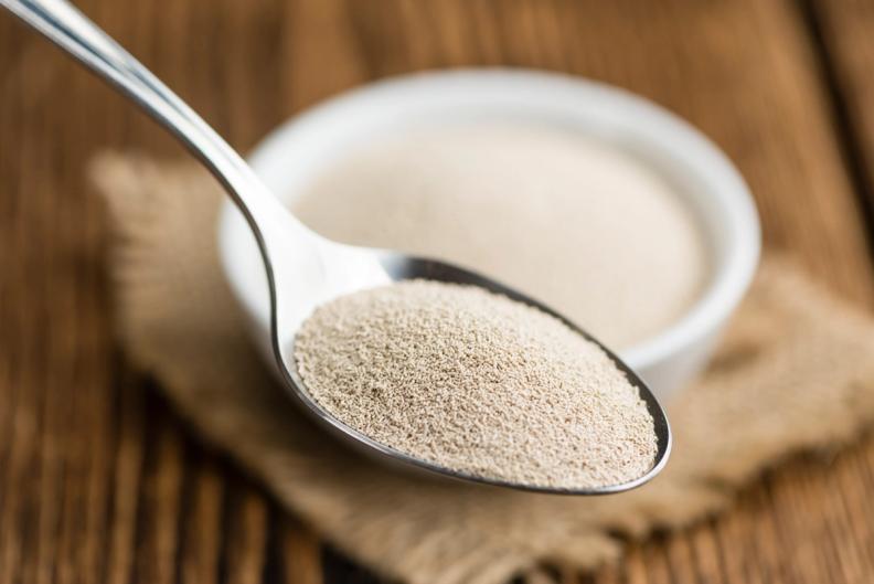 A spoon filled with yeast next to a bowl of yeast on a wooden table.