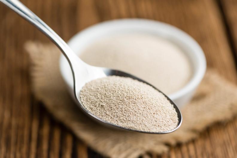 A spoon filled with yeast next to a bowl of yeast on a wooden table.