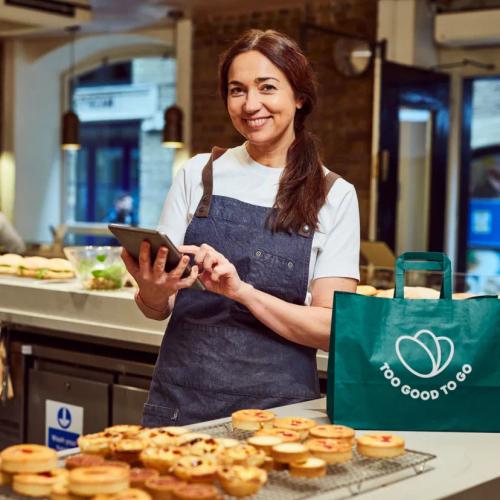 una donna che indossa un grembiule tiene in mano un tablet dietro un bancone che espone prodotti da forno e una borsa verde con la scritta Too Good To Go