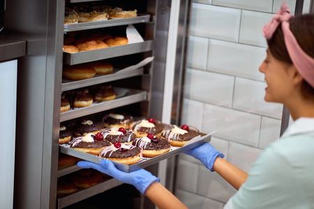 A woman in blue gloves places a tray of chocolate and cherry-topped donuts into a metal bakery rack.