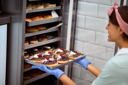 A woman in blue gloves places a tray of chocolate and cherry-topped donuts into a metal bakery rack.