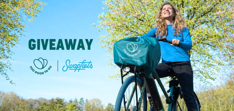 Smiling woman on a bicycle with a 'Too Good To Go' branded basket cover in a sunny park. 'GIVEAWAY' text, Too Good To Go and Swapfiets logos are visible.
