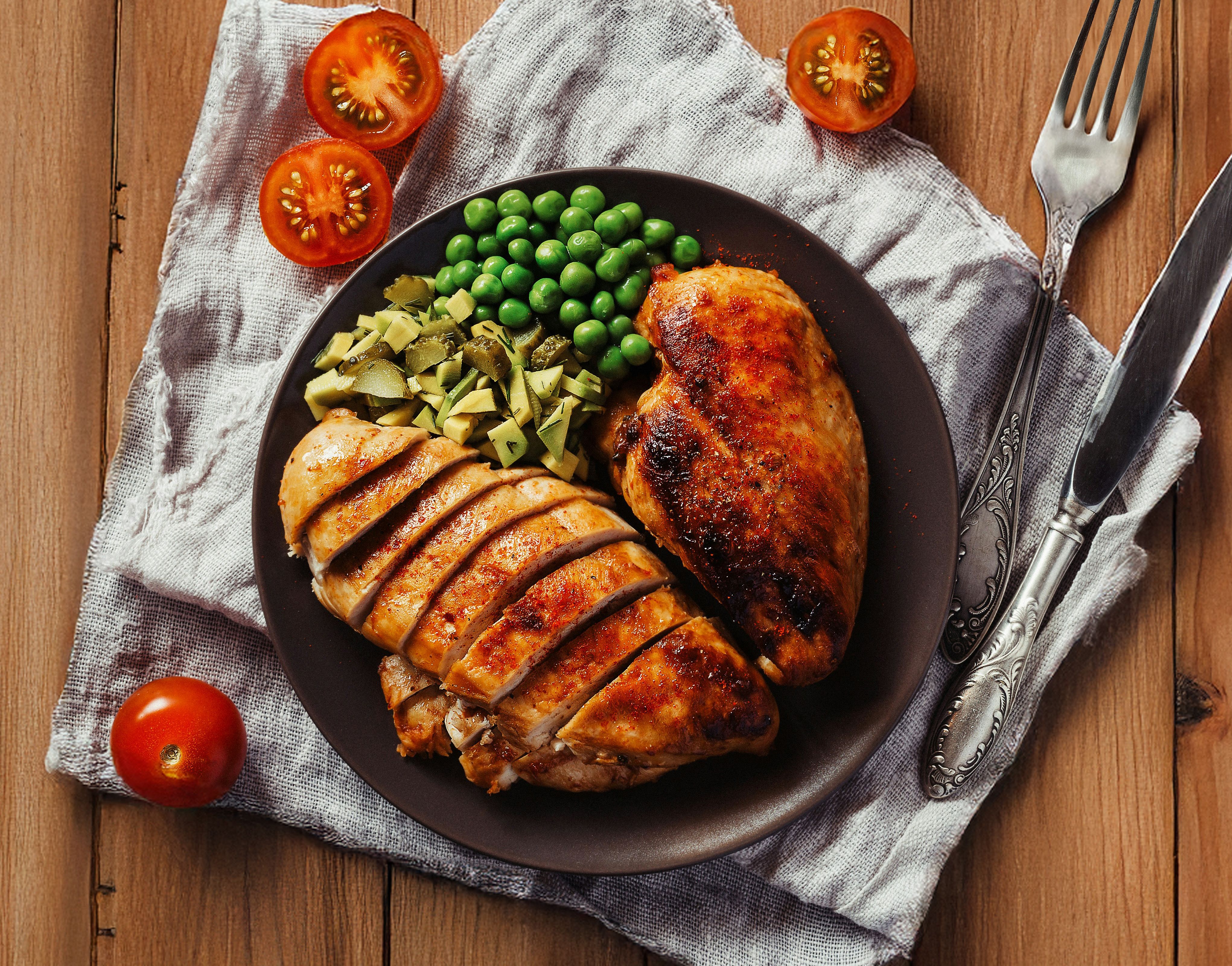 a plate of chicken breast with peas , avocado and tomatoes on a wooden table .