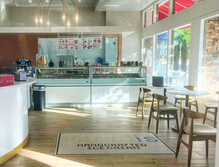 Interior of a bright ice cream shop with display cases, a white counter, and a seating area by large, sunny windows.