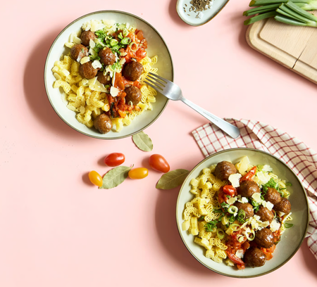 Two bowls of pasta with meatballs, tomato sauce, green onions, and cheese on a pink surface.