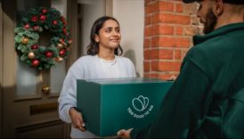 A smiling woman receives a green Too Good To Go box from a delivery person at a doorway with a Christmas wreath.
