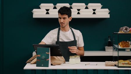A man in a dark apron works at a cafe counter, preparing takeout orders with bags, jars, and a tablet, with takeout containers on a shelf above.