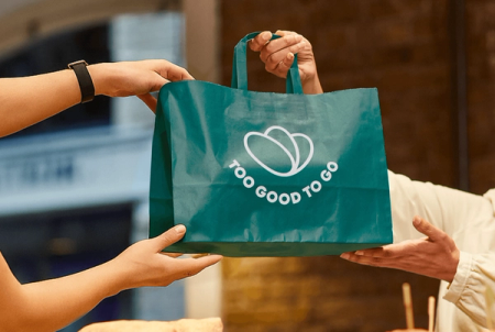 A teal "Too Good To Go" bag being handed between two people.