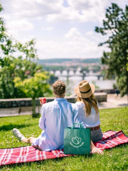 a man and woman sitting on a blanket with a bag that says too good to go