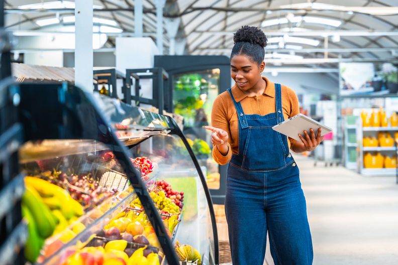 A smiling Black woman in denim overalls inspects colorful fruit in a refrigerated display case, holding a tablet.