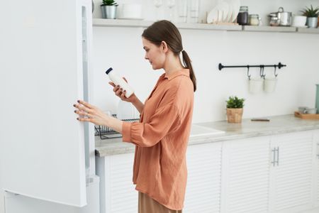 A woman holds a white bottle while opening a refrigerator in a modern kitchen.