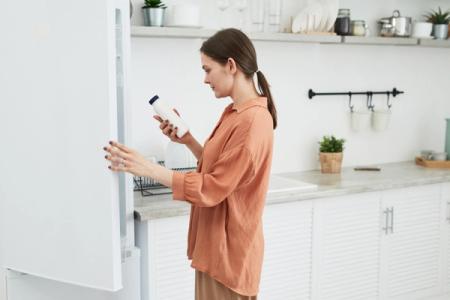 A woman holds a white bottle while opening a refrigerator in a modern kitchen.