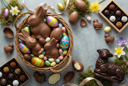An Easter spread featuring a basket of chocolate bunnies and eggs, alongside chocolate lamb, ducklings, truffles, and spring flowers.