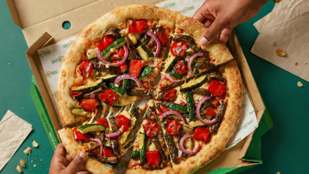 A colorful vegetable-topped pizza in a box with two hands taking slices.