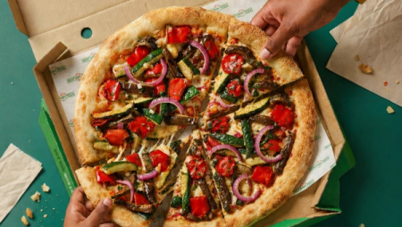 A colorful vegetable-topped pizza in a box with two hands taking slices.