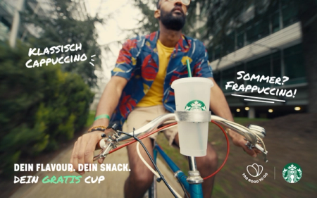 A man cycles outdoors with a Starbucks reusable cup in a bike holder, promoting "Classic Cappuccino," "Summer Frappuccino," and "Your Free Cup" alongside Starbucks and Too Good To Go logos.