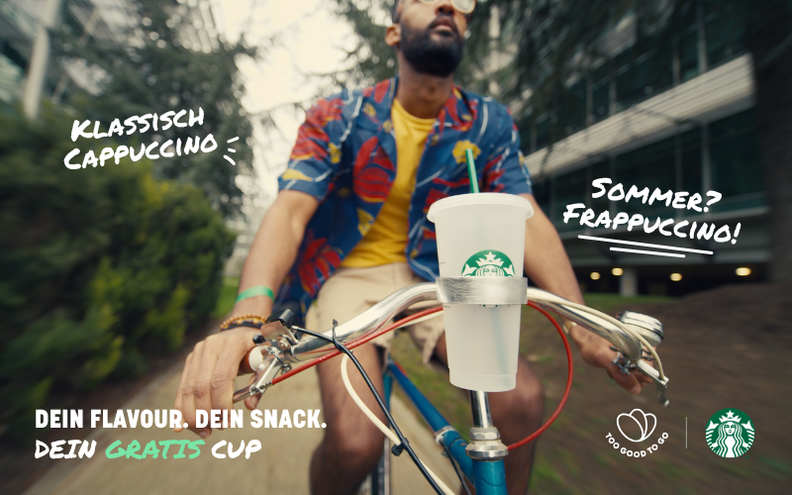 A man cycles outdoors with a Starbucks reusable cup in a bike holder, promoting "Classic Cappuccino," "Summer Frappuccino," and "Your Free Cup" alongside Starbucks and Too Good To Go logos.