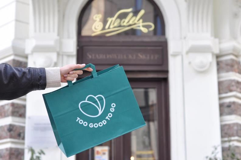 A hand holds a green "Too Good To Go" bag in front of a shop entrance.