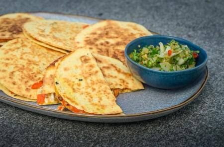 Golden-brown quesadillas filled with red peppers and cheese on a grey plate, served with a blue bowl of green salsa.